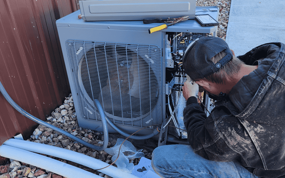 A person repairs an outdoor air conditioning unit, kneeling near its open panel.