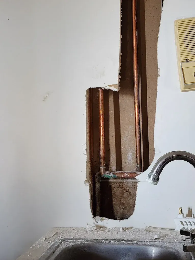 Copper pipes exposed in a wall, near a sink. Water stains, white wall, brown pipes.