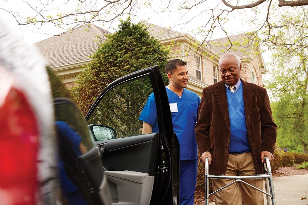 Image of a support worker helping an elderly man with a walker into the passenger seat of a car.
