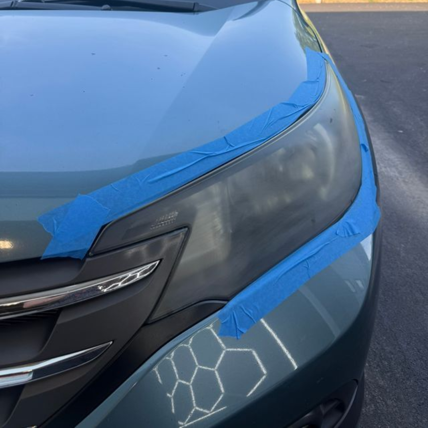 Car headlight taped for restoration, partially hazy against green vehicle.