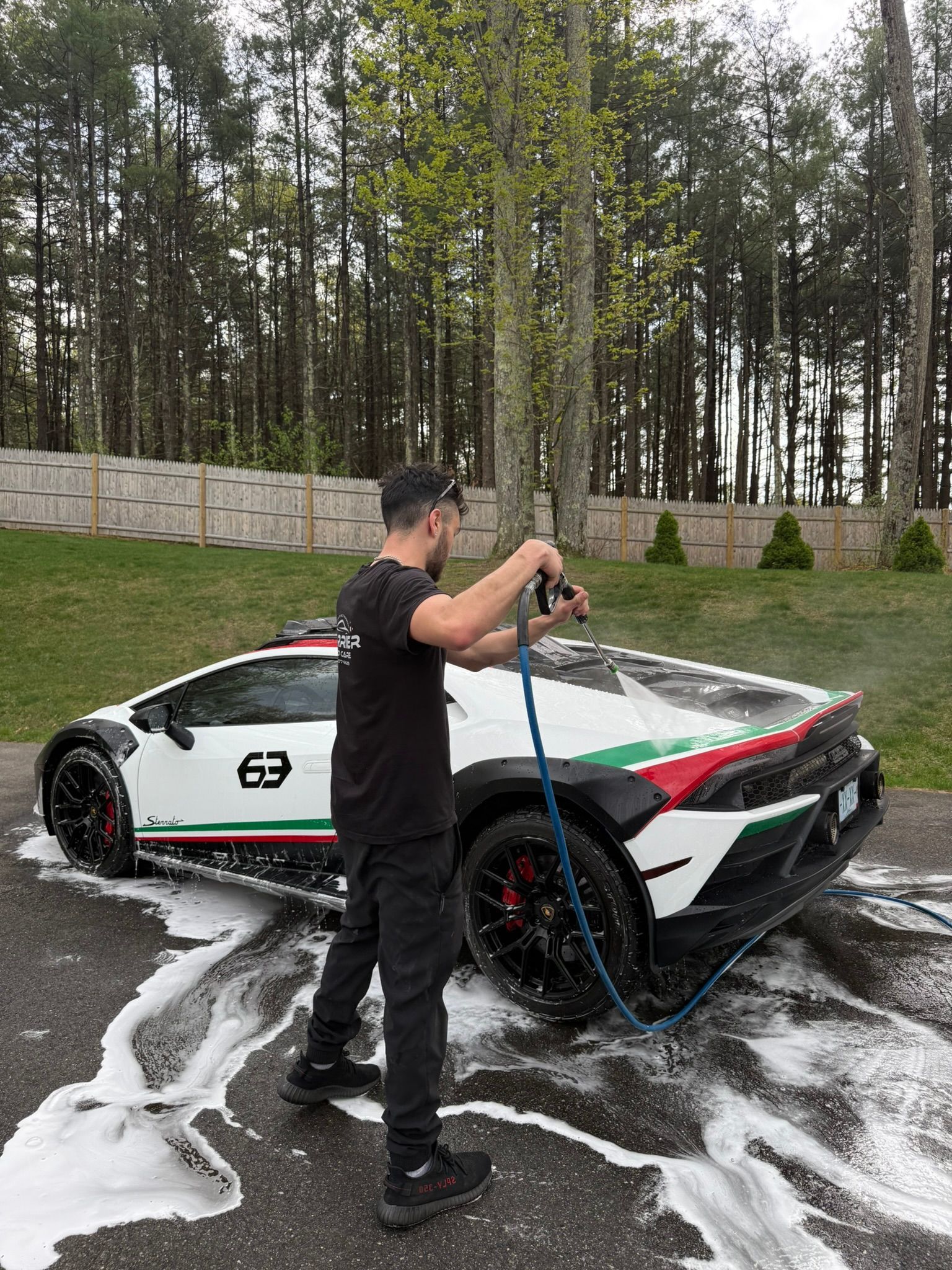 Man washing a white Lamborghini with red and green accents. Outdoors, sudsy, holding a hose.