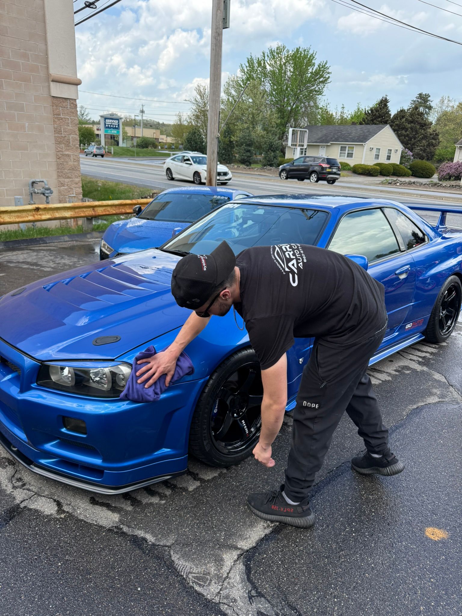 Man washing a blue sports car outdoors; another blue car is visible in the background.