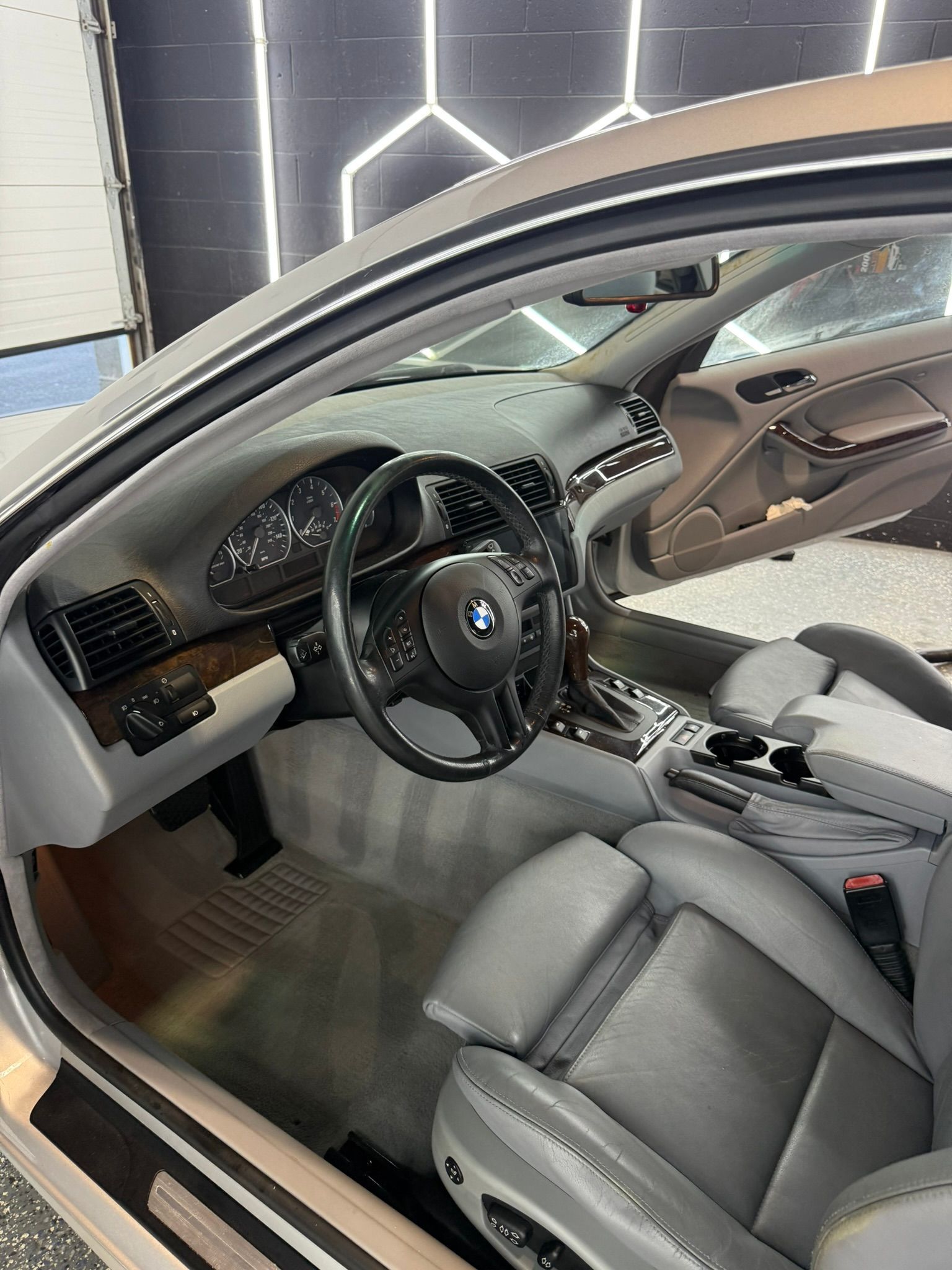 Interior view of a silver BMW.  Black steering wheel, gray leather seats, and wood trim.
