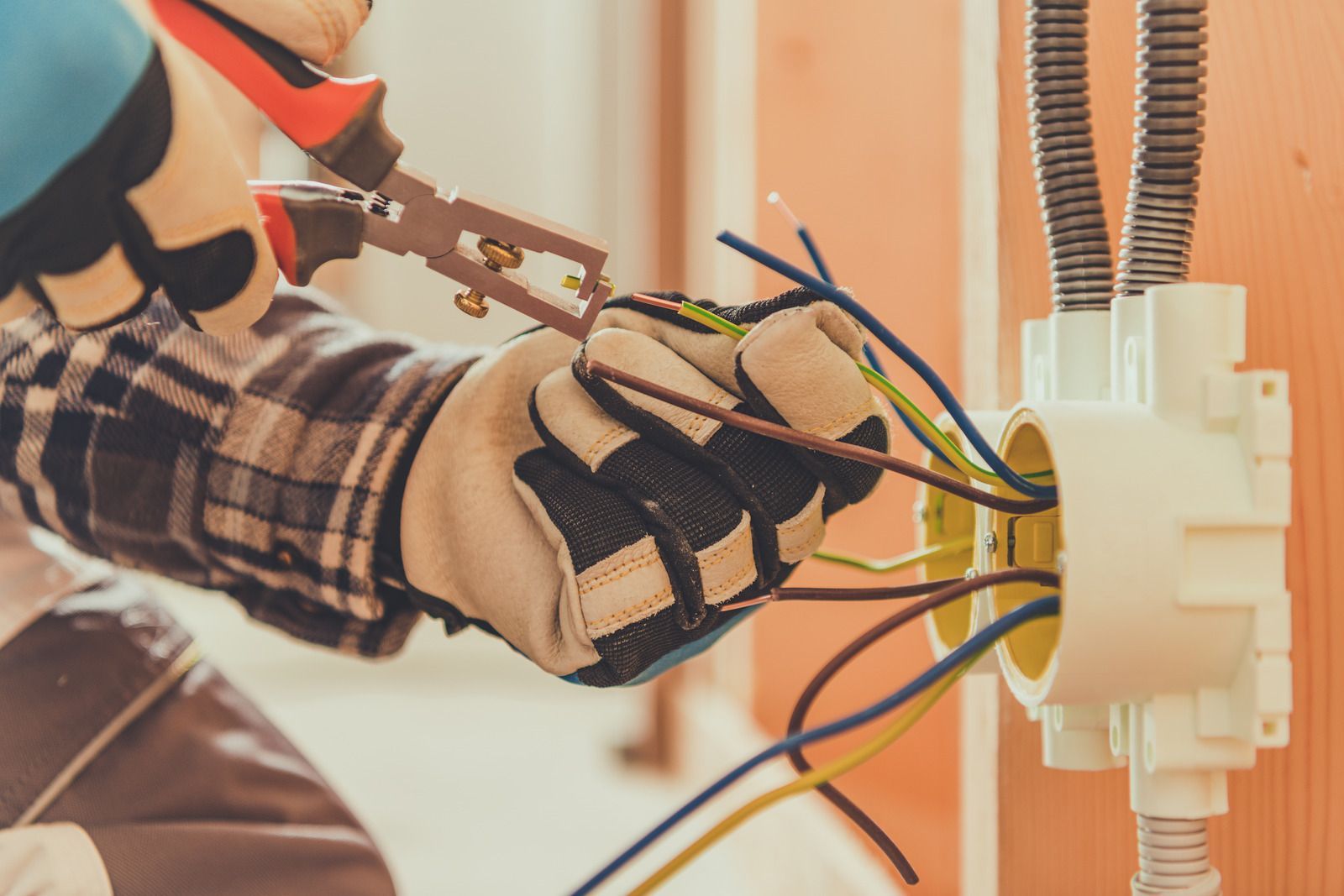 An electrician in work gloves uses wire strippers on electrical cables inside a wall-mounted junction box.