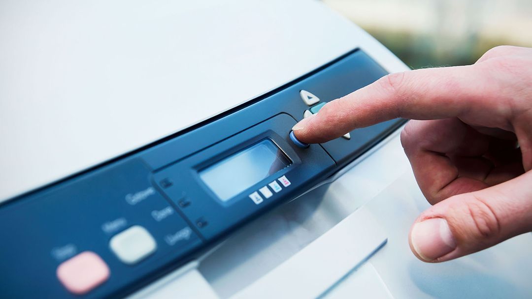 Hand pressing a button on a printer control panel; blue and black with small screen display.