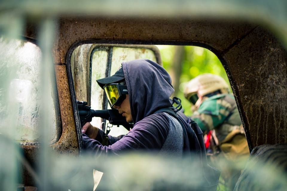 A person in a hoodie and protective goggles aims an airsoft gun from an old truck.