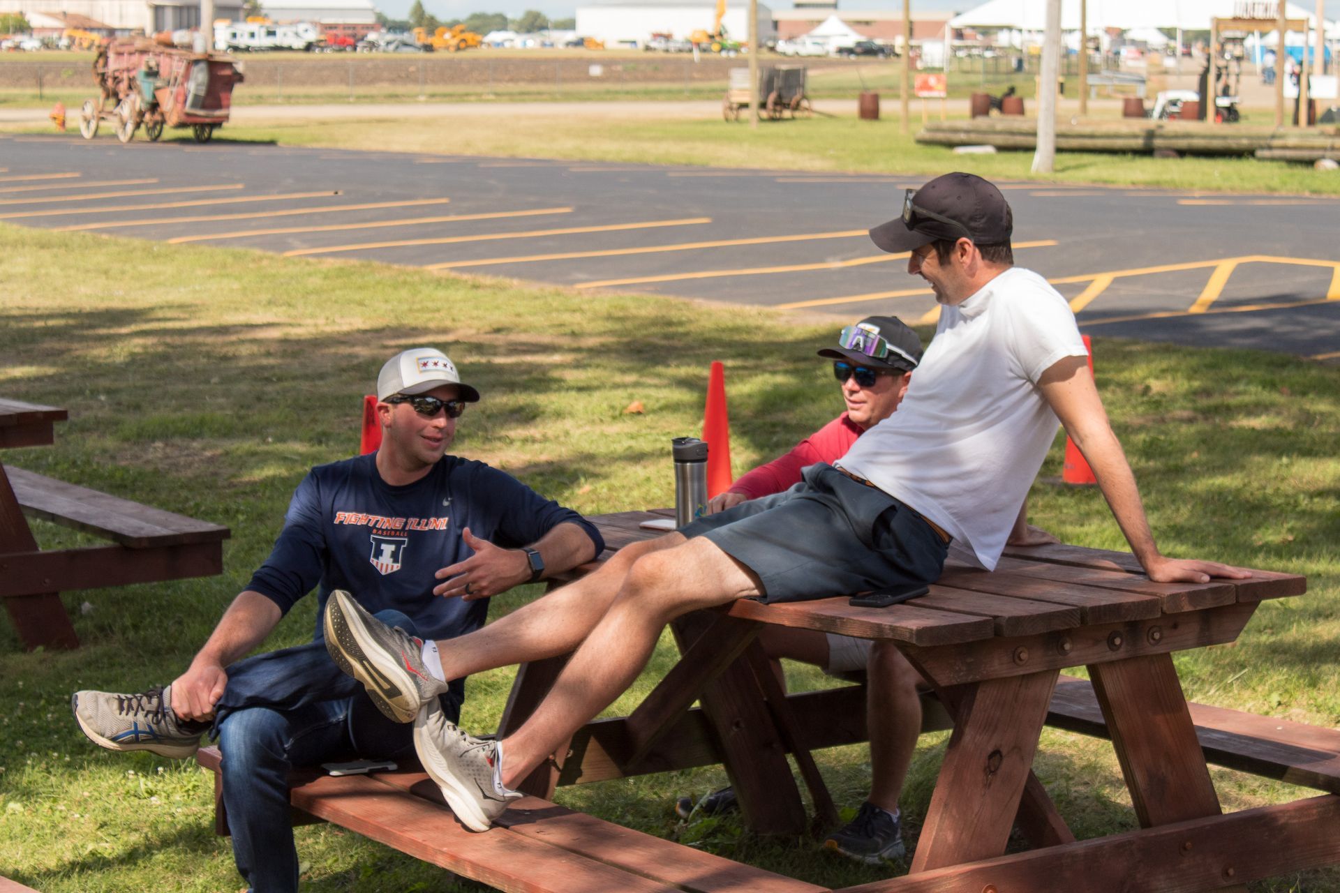 Three men relax on a picnic table, outdoors. One gestures, all wear hats, light and medium skin tones.