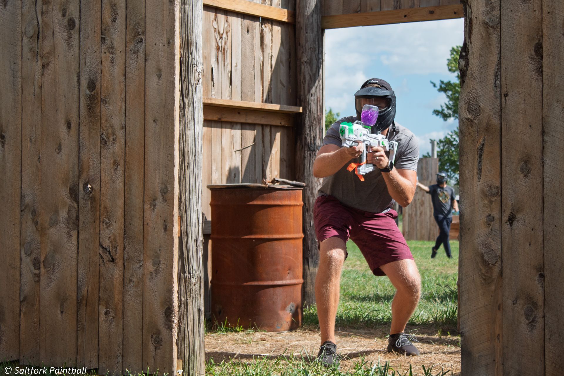 Man with gel blaster peeking out of wooden structure, aiming, wearing protective mask. Outdoors, sunny.