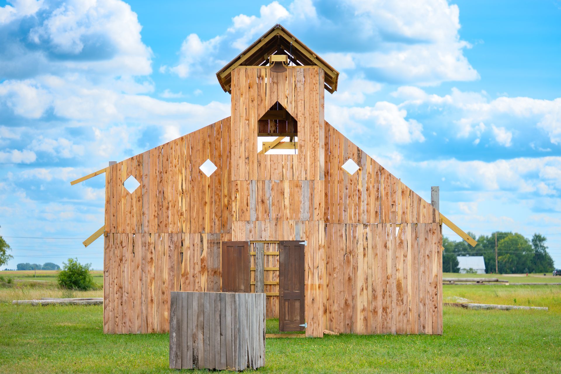Wooden structure under construction in a field. Sunny sky, open doorway, barrel in the foreground.