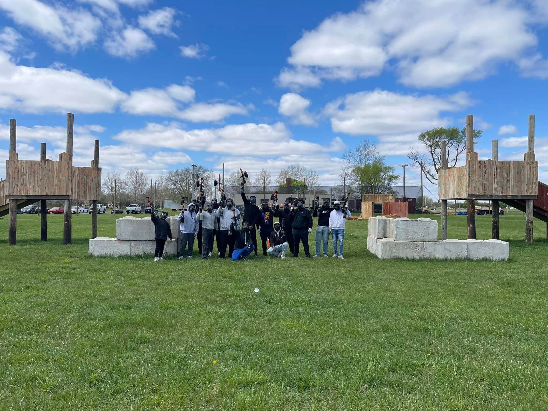 Group of people posing in a grassy field, wooden structures, and concrete blocks. Blue sky with clouds.