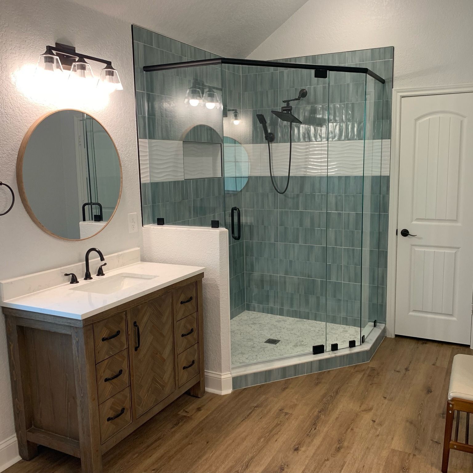 Modern bathroom with wood vanity, round mirror, and glass shower with blue tile accents