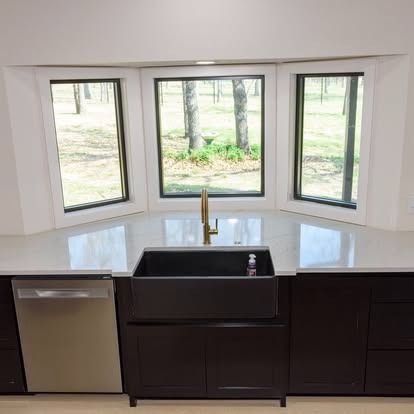 Modern kitchen sink beneath bay windows, with dark cabinets and a stainless-steel dishwasher.