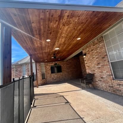 Covered brick patio with wood ceiling, black railing, hanging grill, and concrete floor in sunlight