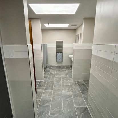Public restroom with gray tile floor, white stalls, and sinks under bright ceiling lights.