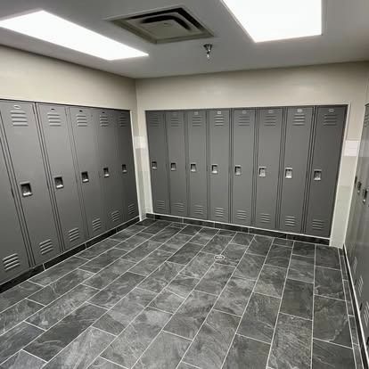 Empty locker room with gray lockers, tiled floor, and fluorescent ceiling lights.