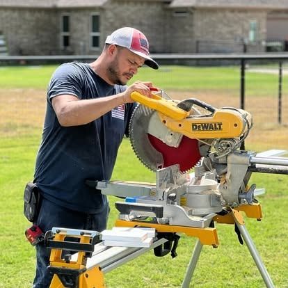 Worker using a DeWalt miter saw outdoors on a folding stand, cutting wood on a grassy lawn.