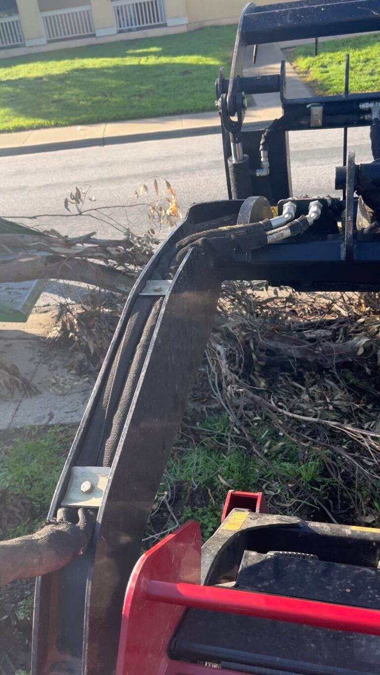 A loader lifting debris near a street and grass, with the machine's arm and bucket visible.