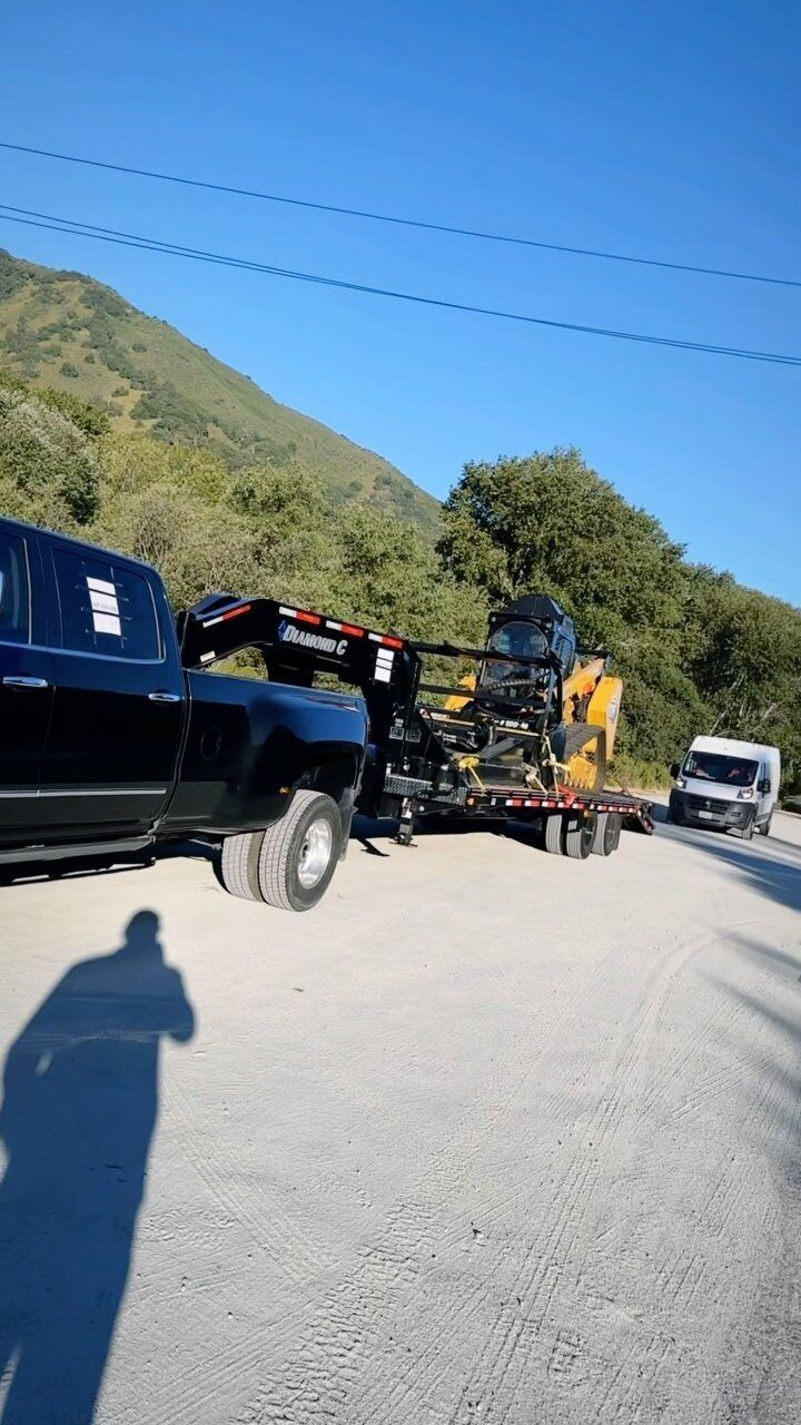 A black pickup truck towing heavy machinery on a trailer on a gravel road near a mountain.