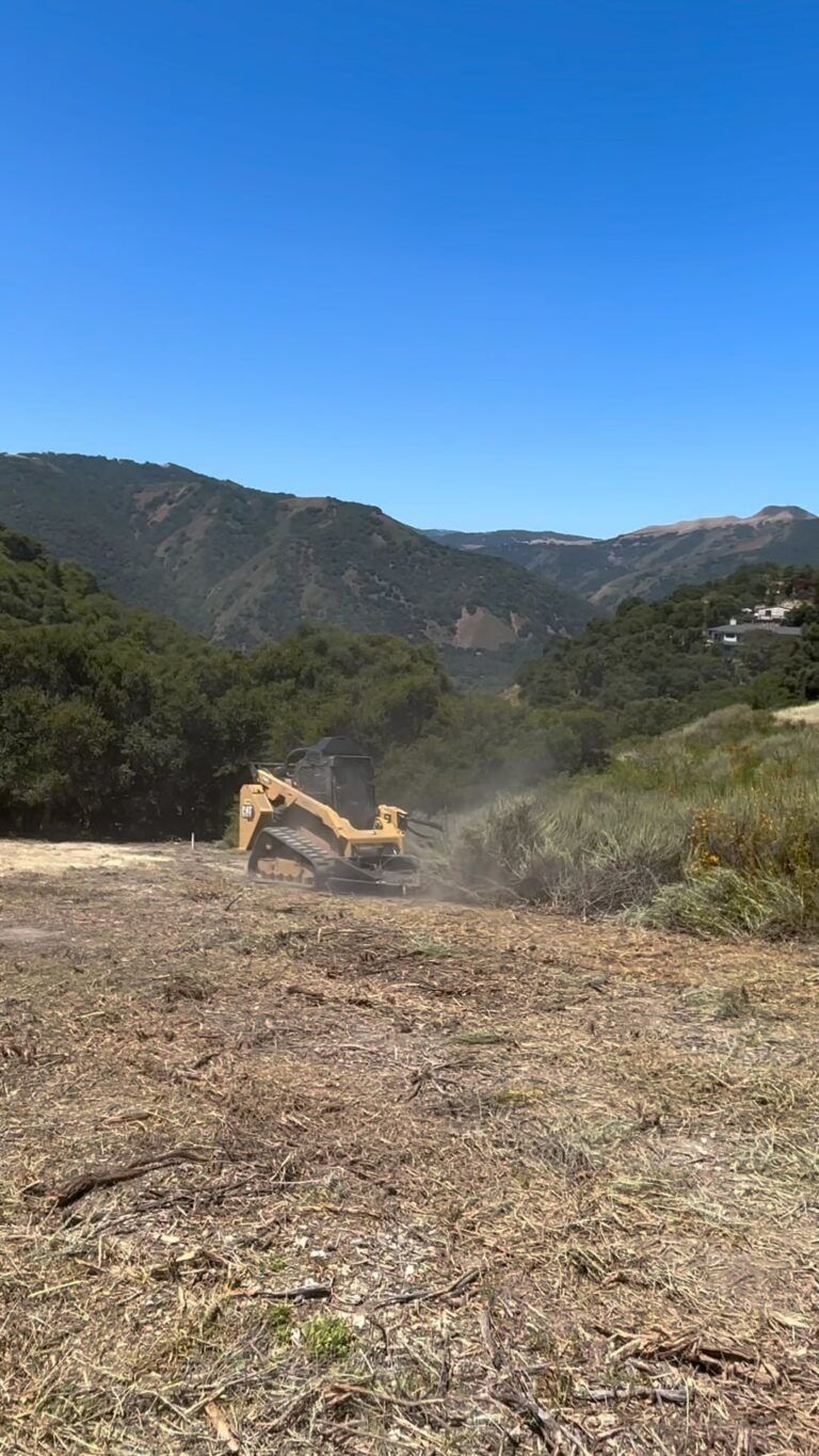 A yellow excavator clearing brush on a hillside with a view of rolling hills under a blue sky.