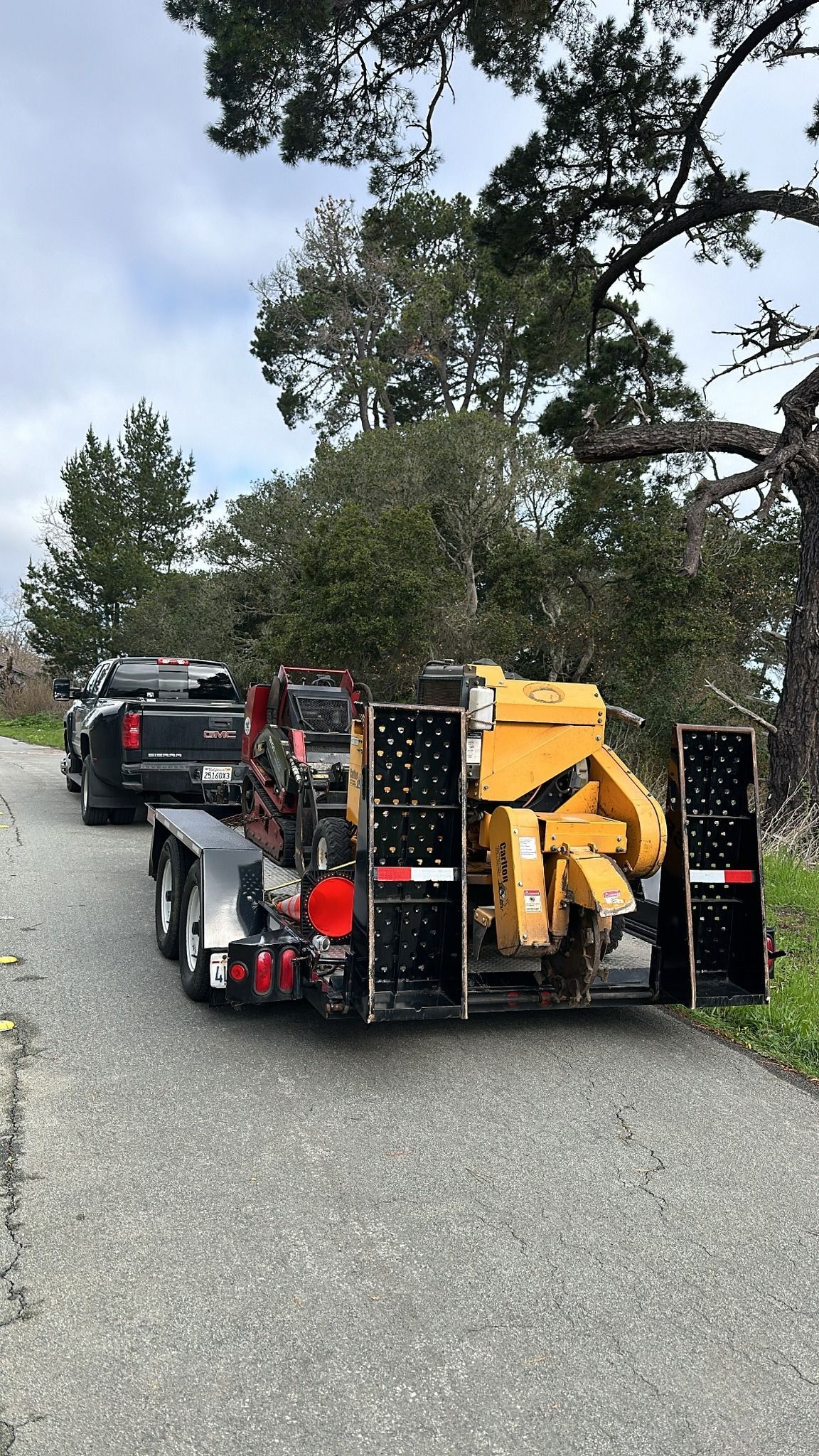 Truck pulling a trailer with a yellow wood chipper on a paved road. Trees in background, cloudy sky.