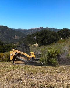 Yellow tracked brush cutter clearing dry brush on a hillside with mountains in the background.