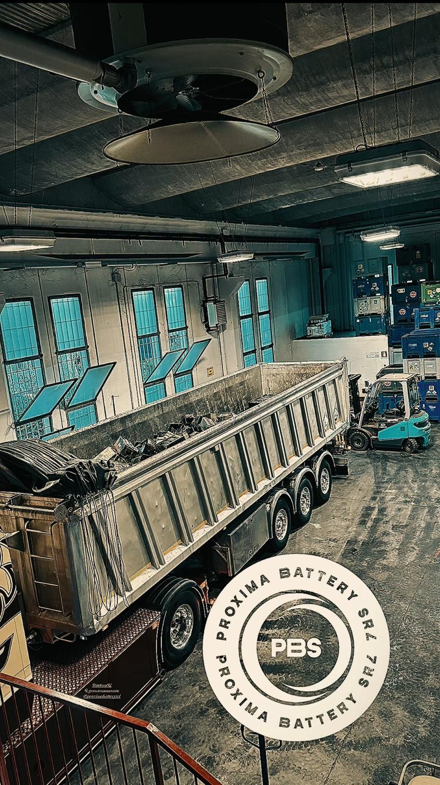Uno spazio industriale con un grande rimorchio ribaltabile argentato parcheggiato, ventilatori a soffitto e un logo circolare sovrapposto di Proxima Battery.