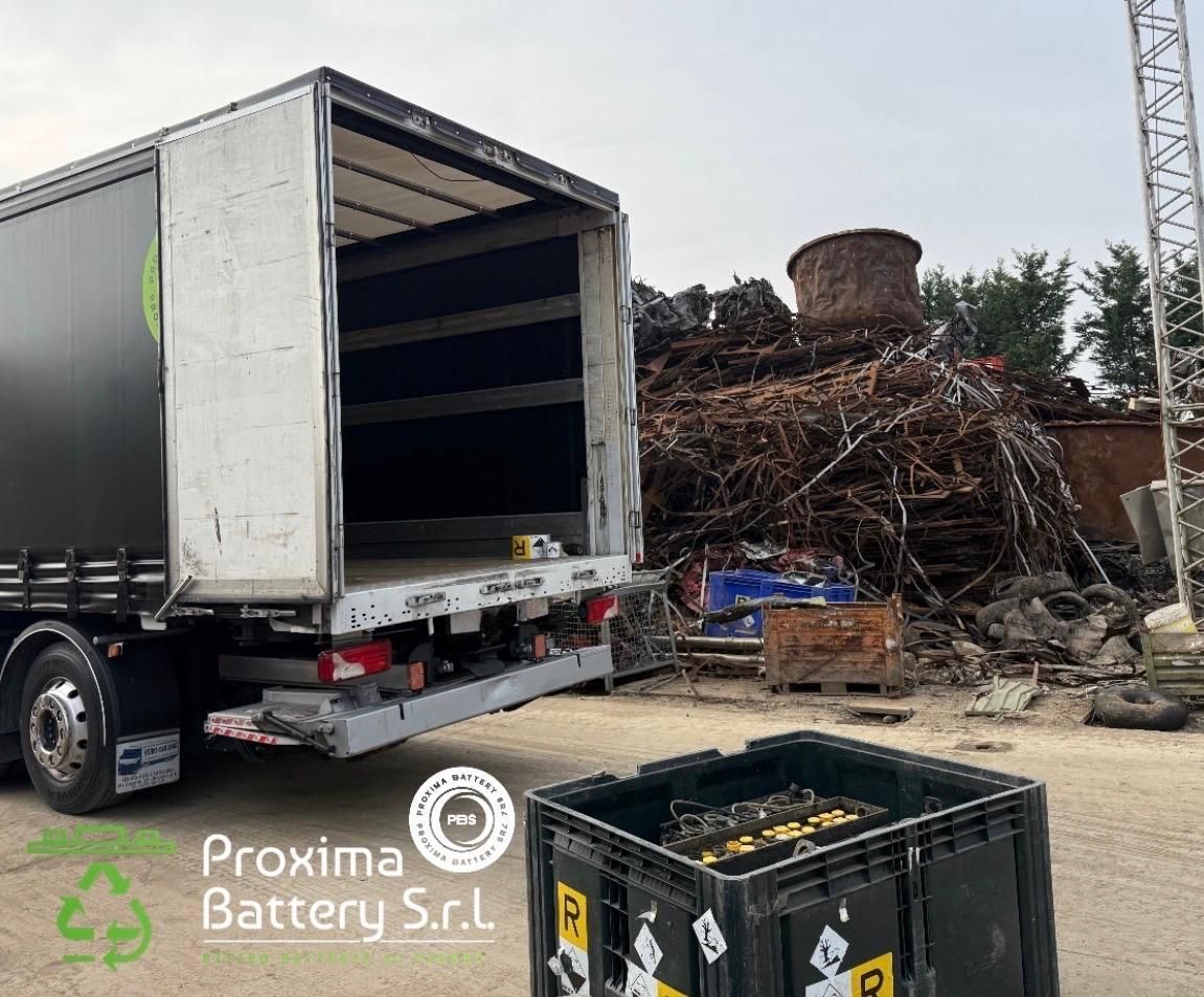 Un camion parcheggiato accanto a un grande cumulo di rottami metallici sotto un sole splendente, con il logo di Proxima Battery Srl in basso.