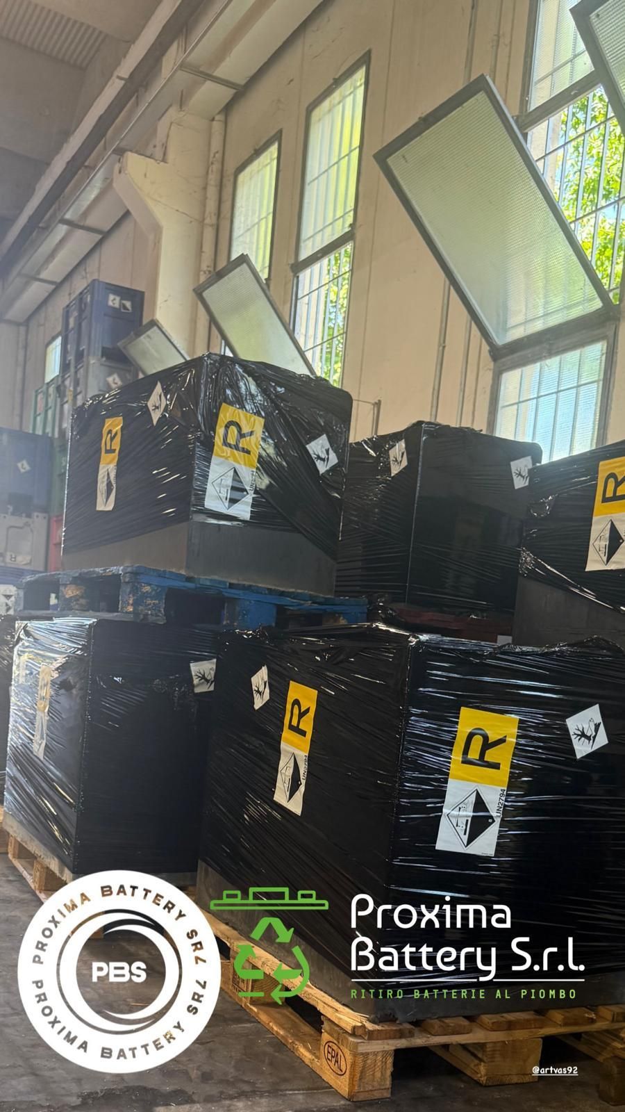 All'interno di un magazzino, si vedono pallet di legno impilati e avvolti in plastica nera con il marchio Proxima Battery Srl.