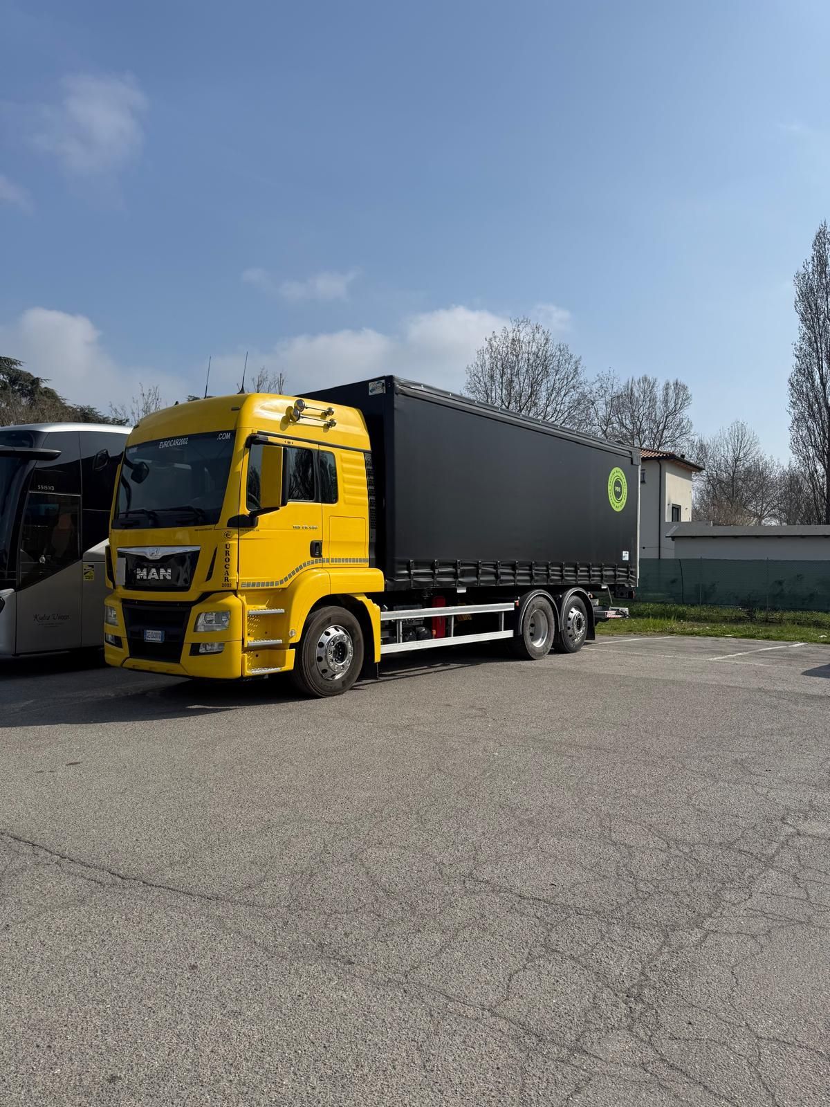 Un camion MAN giallo con rimorchio nero parcheggiato su un piazzale ghiaioso sotto un cielo azzurro e limpido.