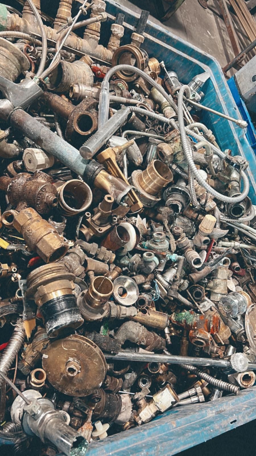 Un bidone blu pieno di un mucchio disordinato di vari raccordi idraulici in ottone, tubi e rottami metallici.