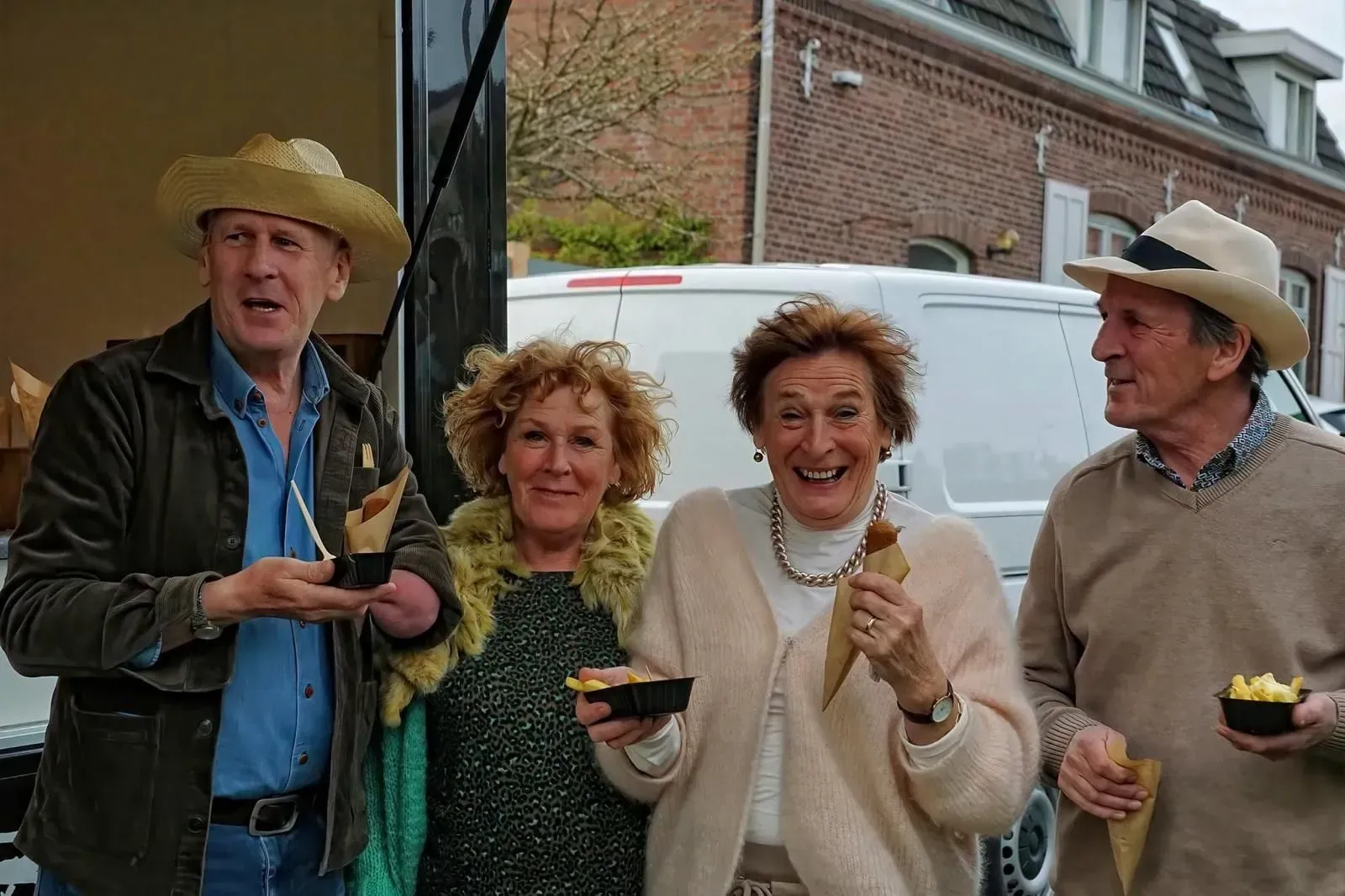 Vier lächelnde Menschen halten Essen in der Hand, im Freien. Zwei Frauen und zwei Männer, alle tragen Hüte, genießen Snacks.
