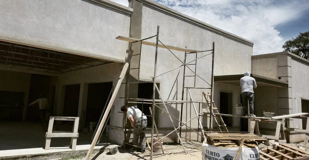 Obreros de la construcción enyesando el exterior de un edificio de color claro, utilizando andamios en un día soleado.
