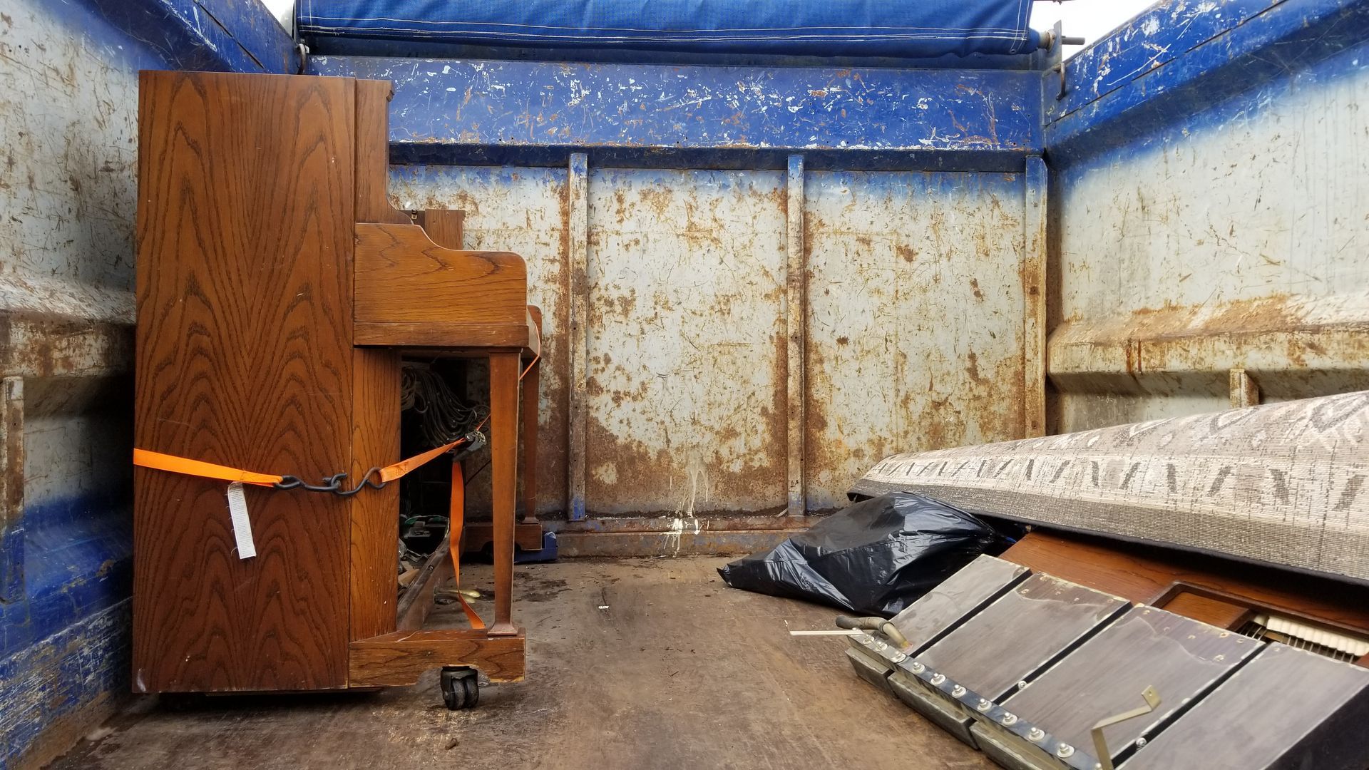 A wooden upright piano secured with a strap sits inside the weathered cargo area of a truck with a mattress nearby.
