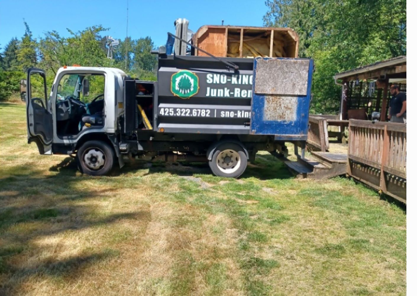 A white junk removal truck parked on a grass lawn next to a wooden deck, with a filled cargo bed visible.