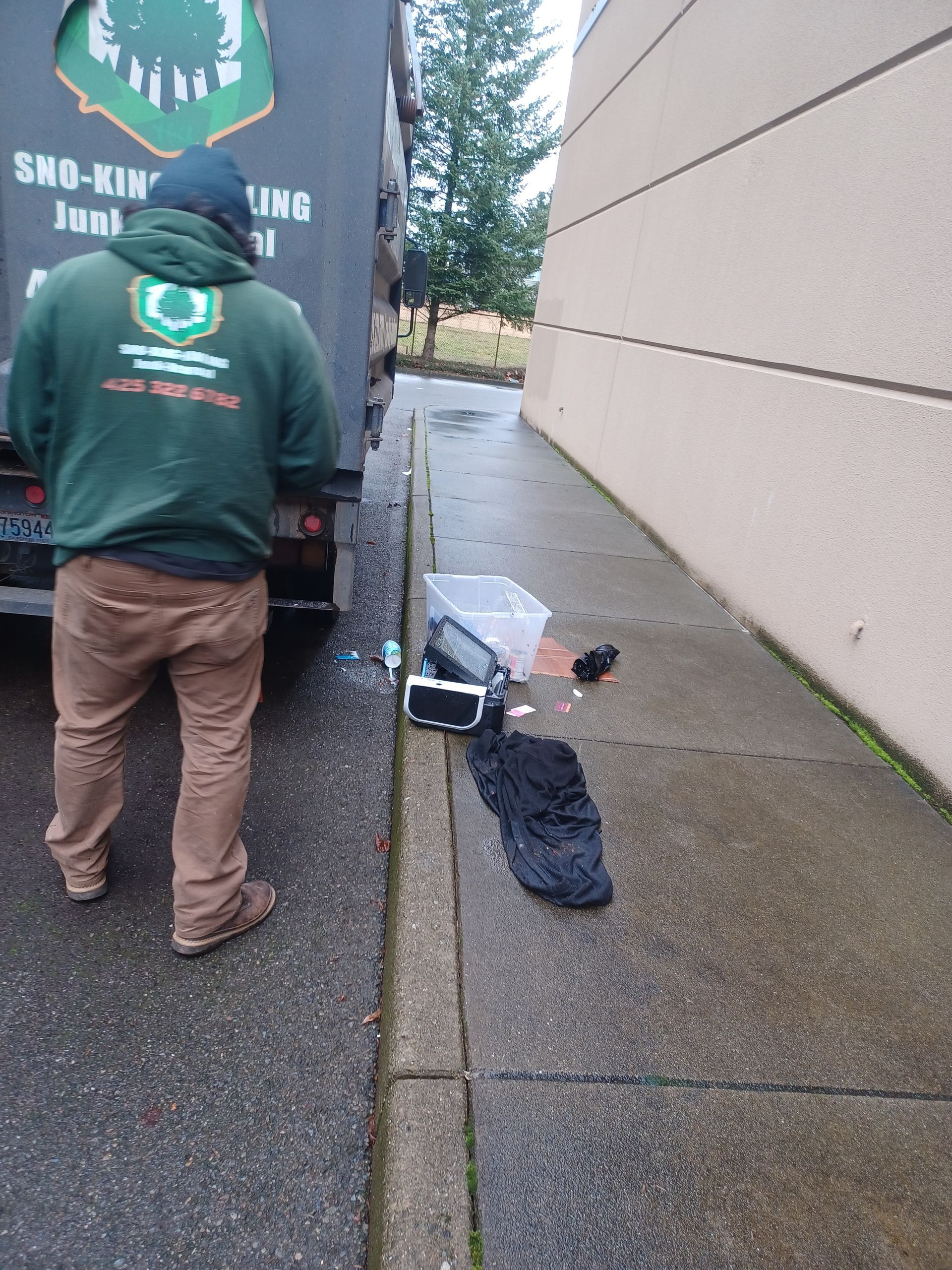 A worker in a green hoodie stands by a junk removal truck on a sidewalk next to a plastic bin and miscellaneous items.