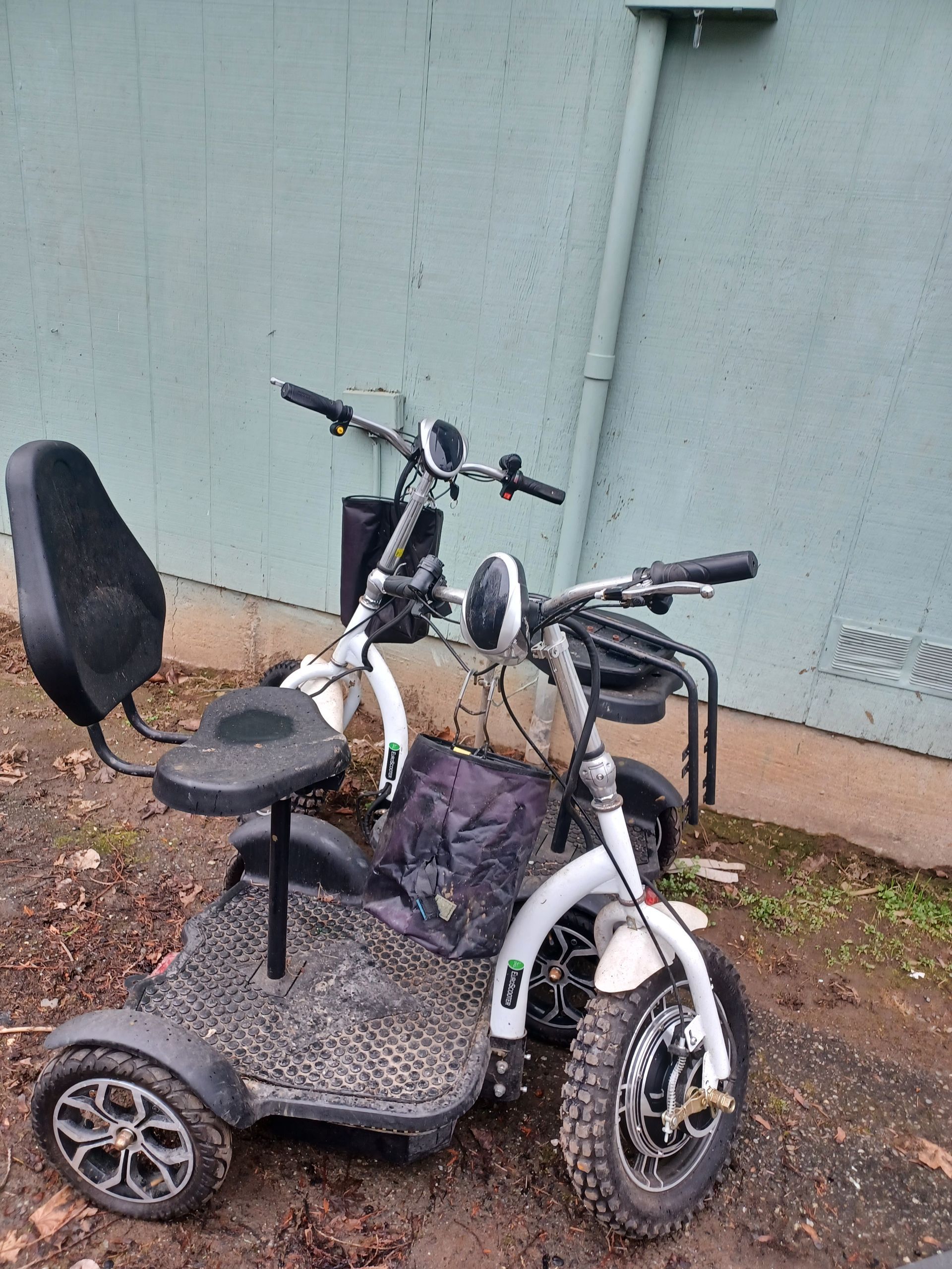 A white, three-wheeled mobility scooter with a black seat and handlebars, parked on dirt near a light green wall.