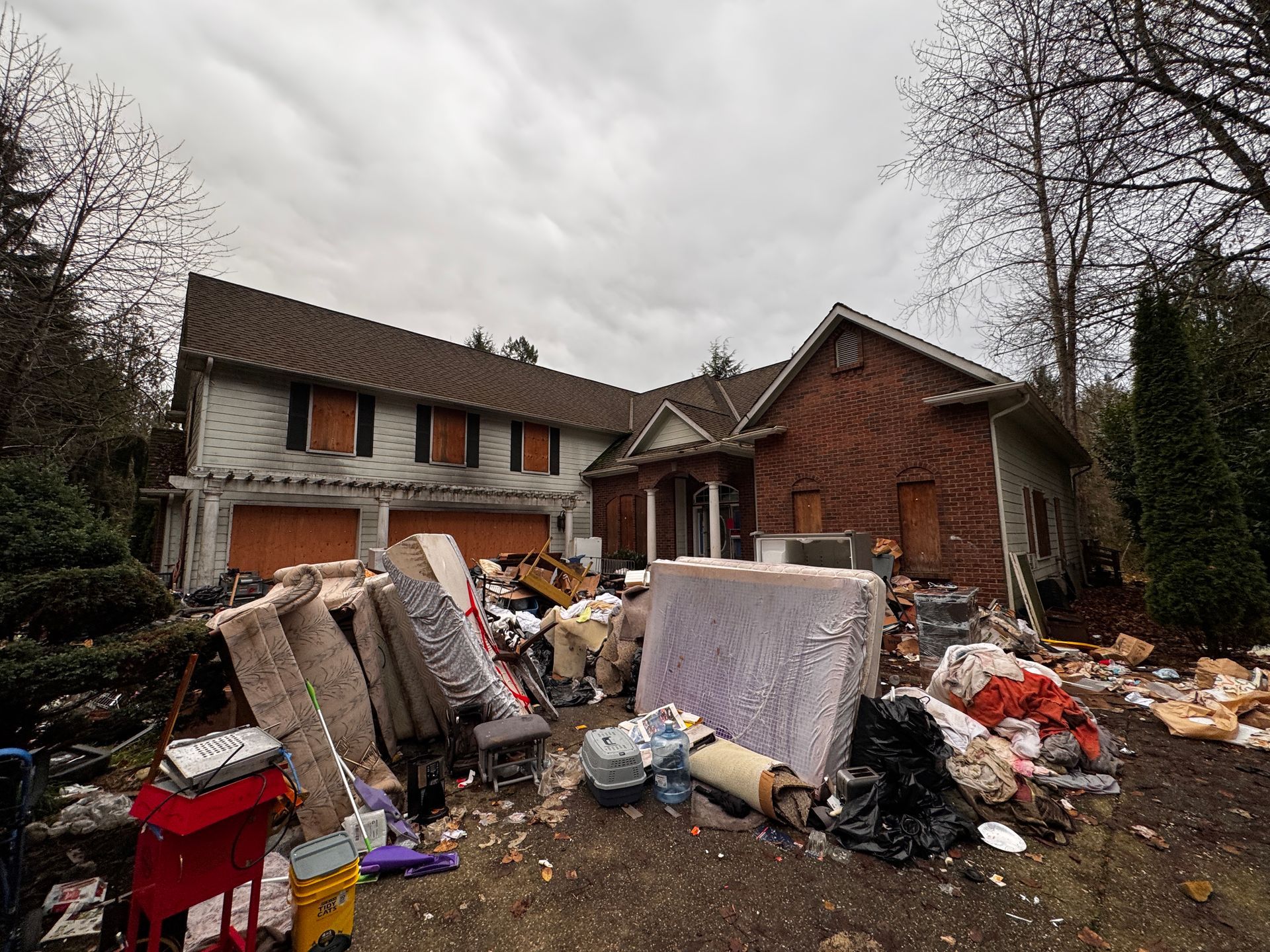 A two-story house with boarded windows sits behind a large, cluttered pile of mattresses, trash, and discarded items.