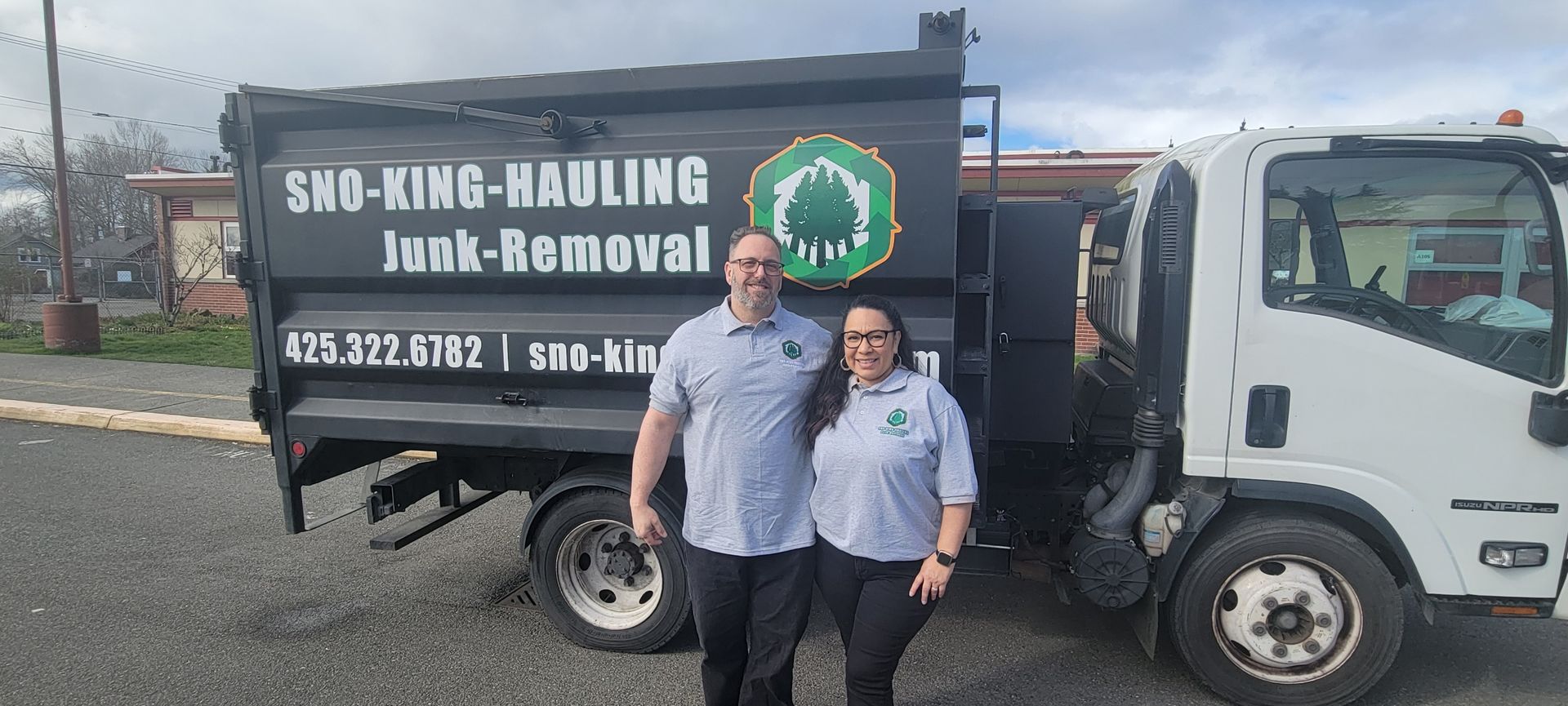 Two people stand smiling in front of a black Sno-King Hauling junk removal truck parked on a paved lot.