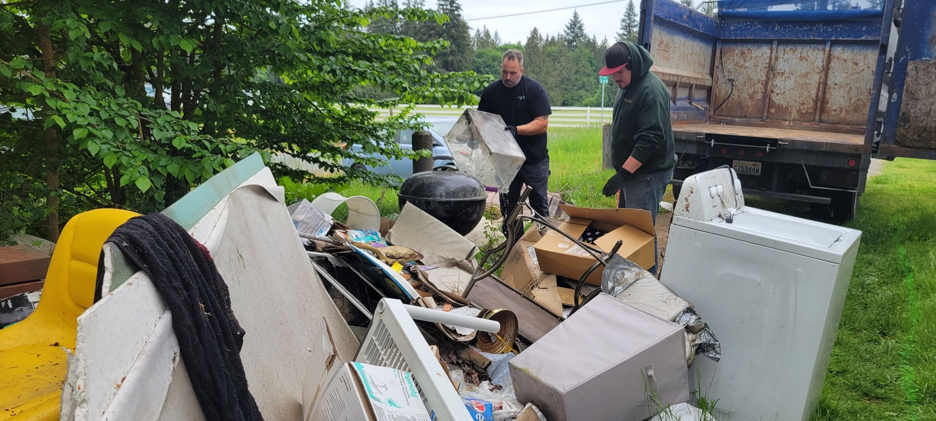 Two people load junk, including a white appliance and various boxes, into the back of a blue dump truck parked on grass.
