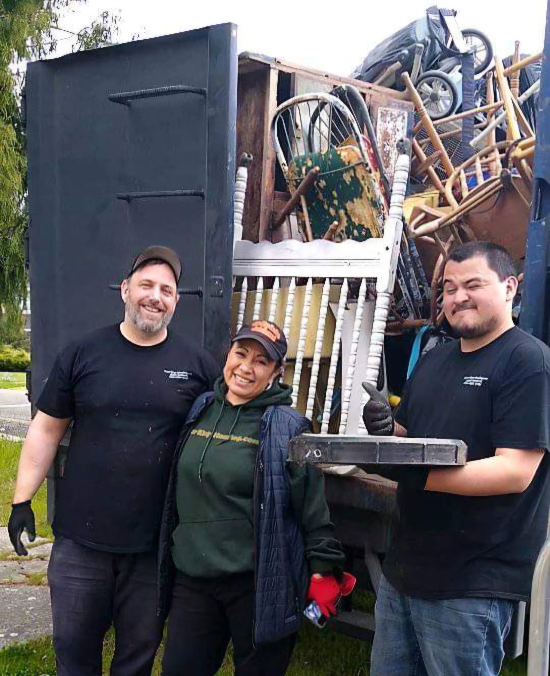 Three people in branded work uniforms smile in front of a truck filled with a variety of furniture and household items.