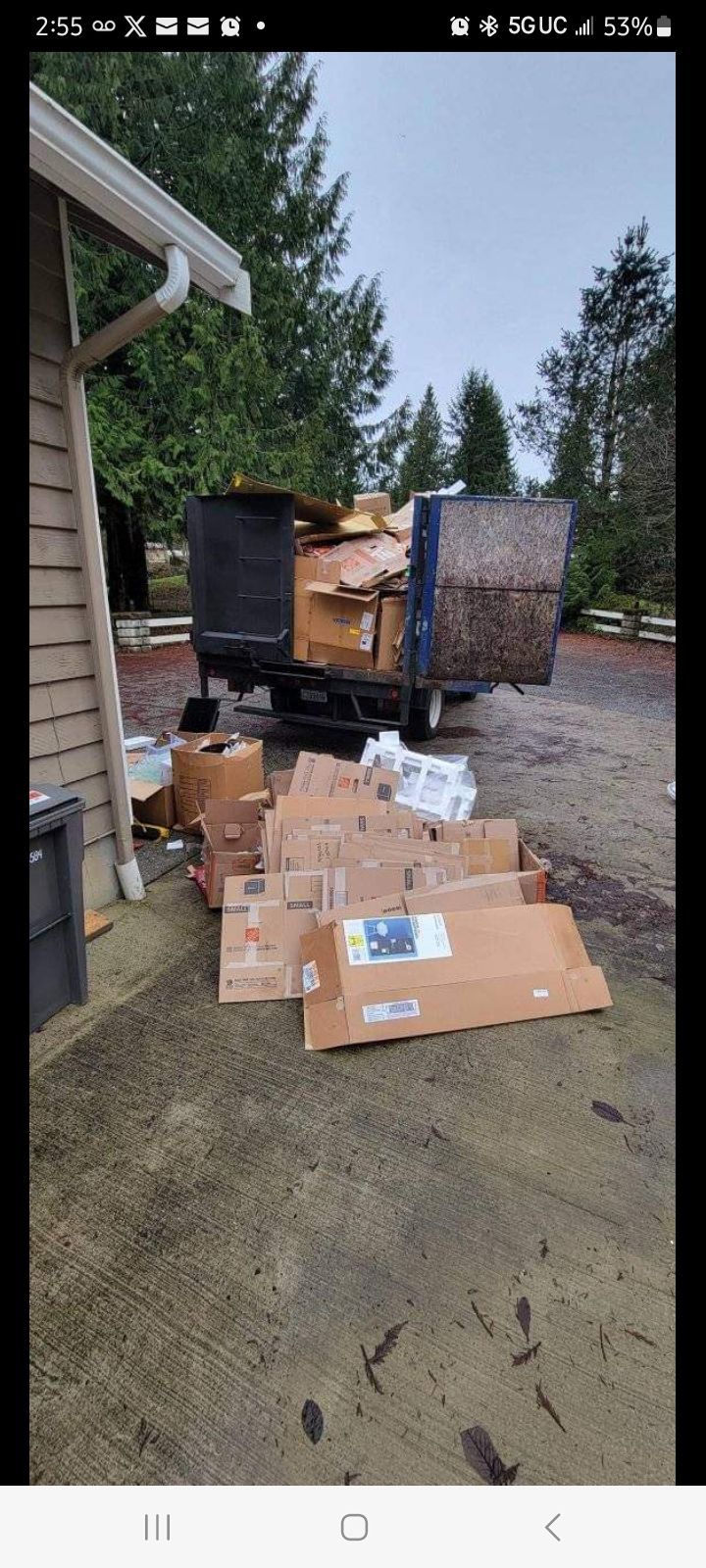 A pile of flattened cardboard boxes sits on a concrete driveway near a house with a loaded trailer parked in the background.