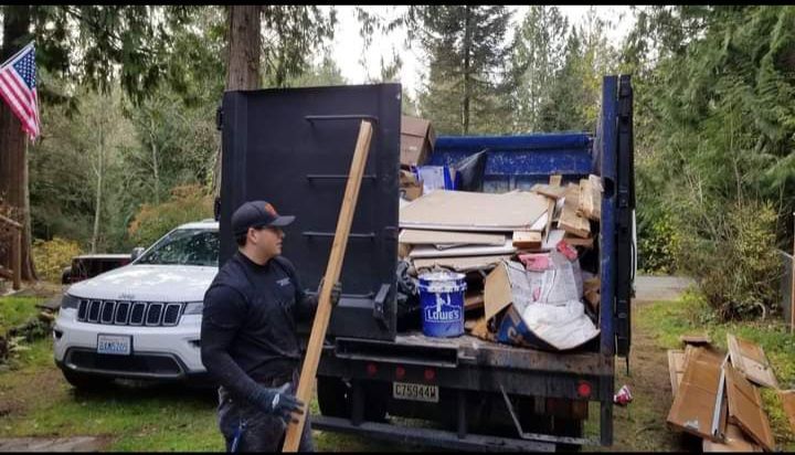 A worker holding a wooden plank stands before a dumpster filled with debris, parked near a white car and a wooded area.
