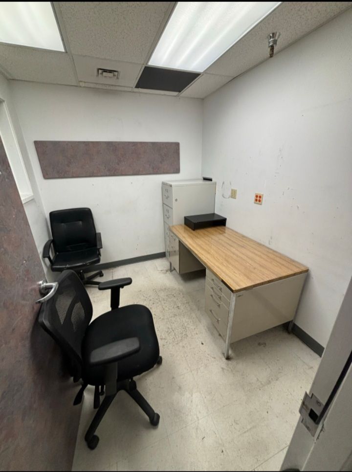 A small, sparse office with two black office chairs, a light-colored metal desk, and a filing cabinet.