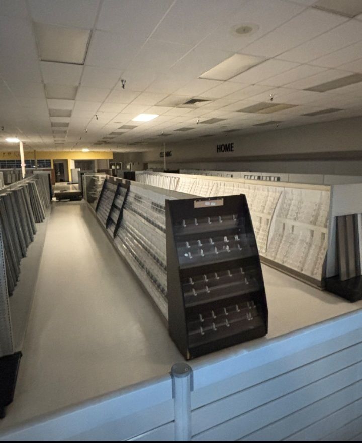 An interior view of a vacant retail space featuring long, empty white shelving units and a single dark display stand.
