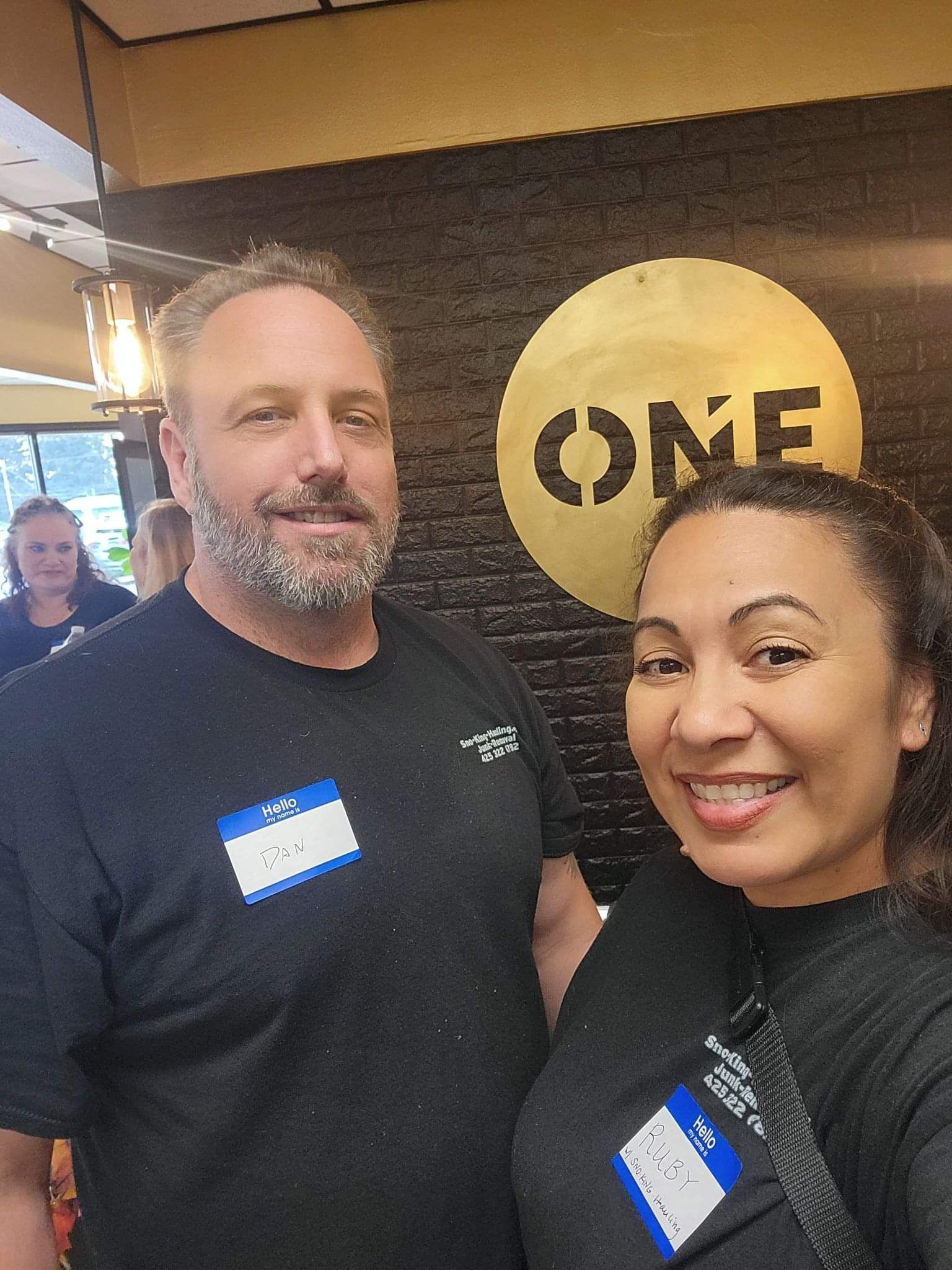 A man and woman wear name tags and black shirts while standing in front of a circular sign that says ONE.
