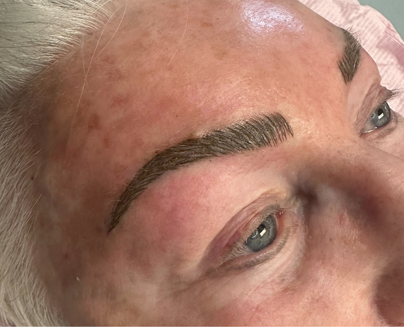 Close-up of a woman's face with freshly tattooed eyebrows, gray hair, and blue eyes.