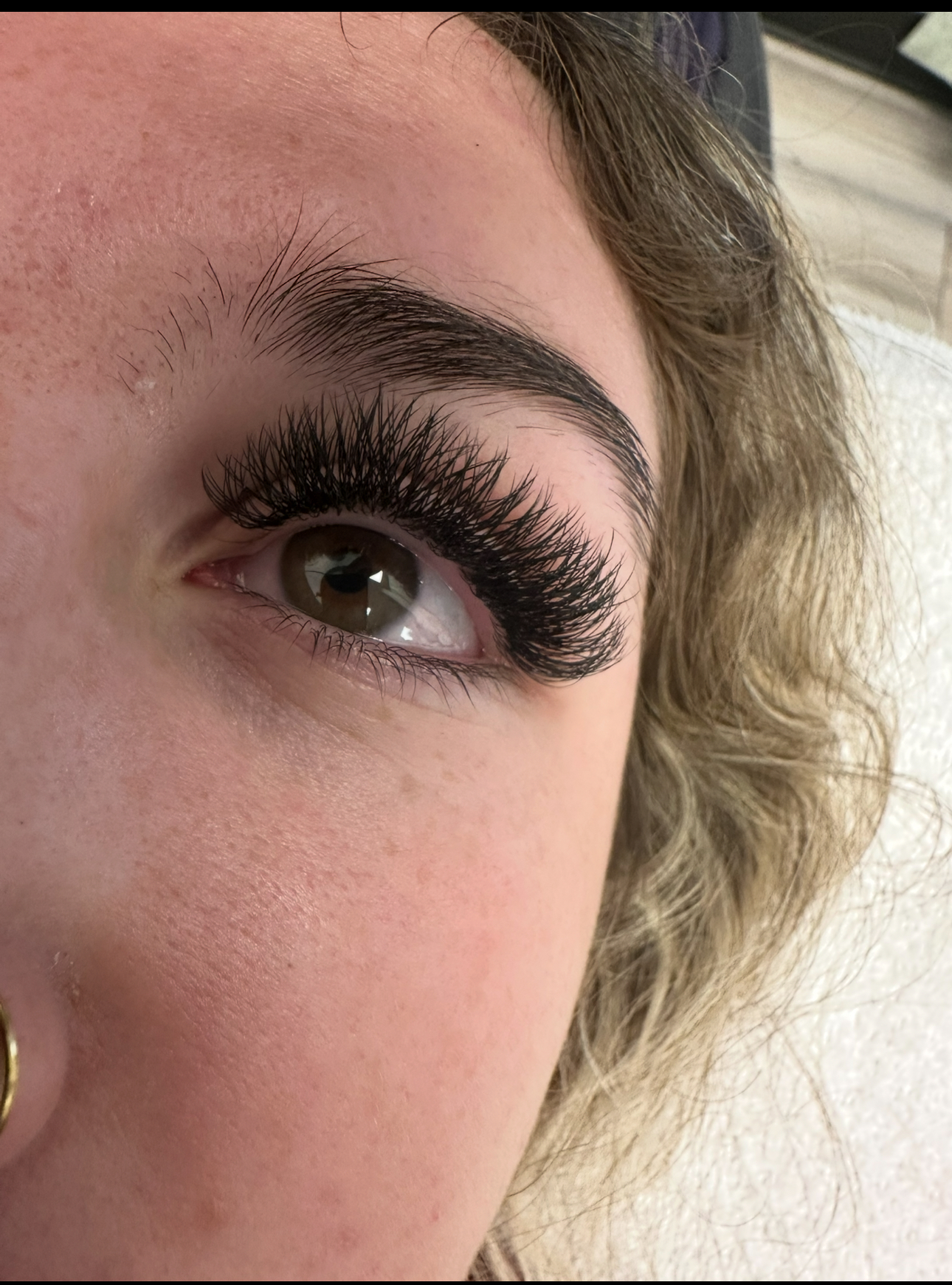 Close-up of an eye with dark, thick eyelash extensions and filled-in eyebrows. Skin is pale, with a nose ring.