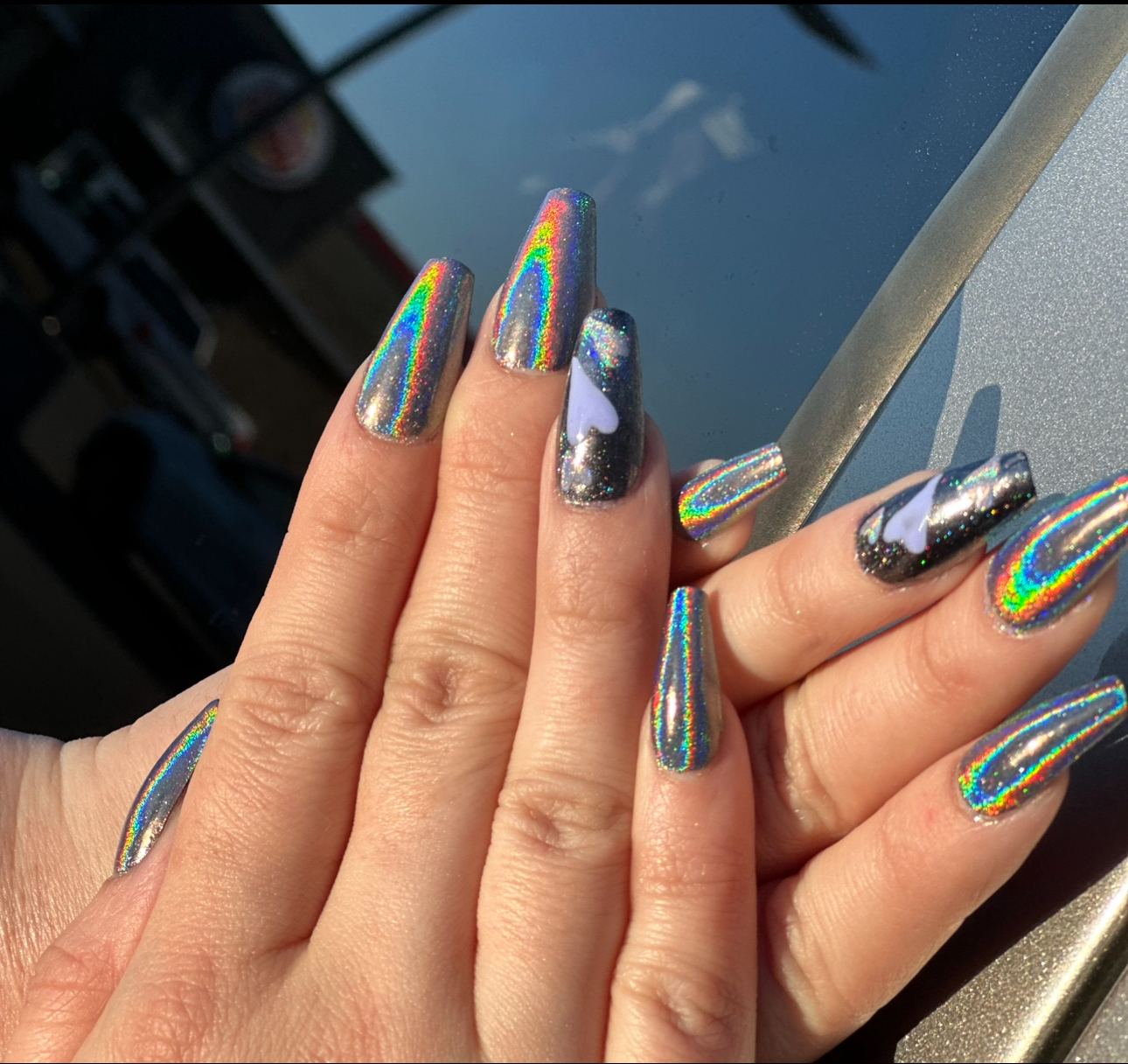 Hands with long, silver holographic nails. One nail is purple and black with glitter.