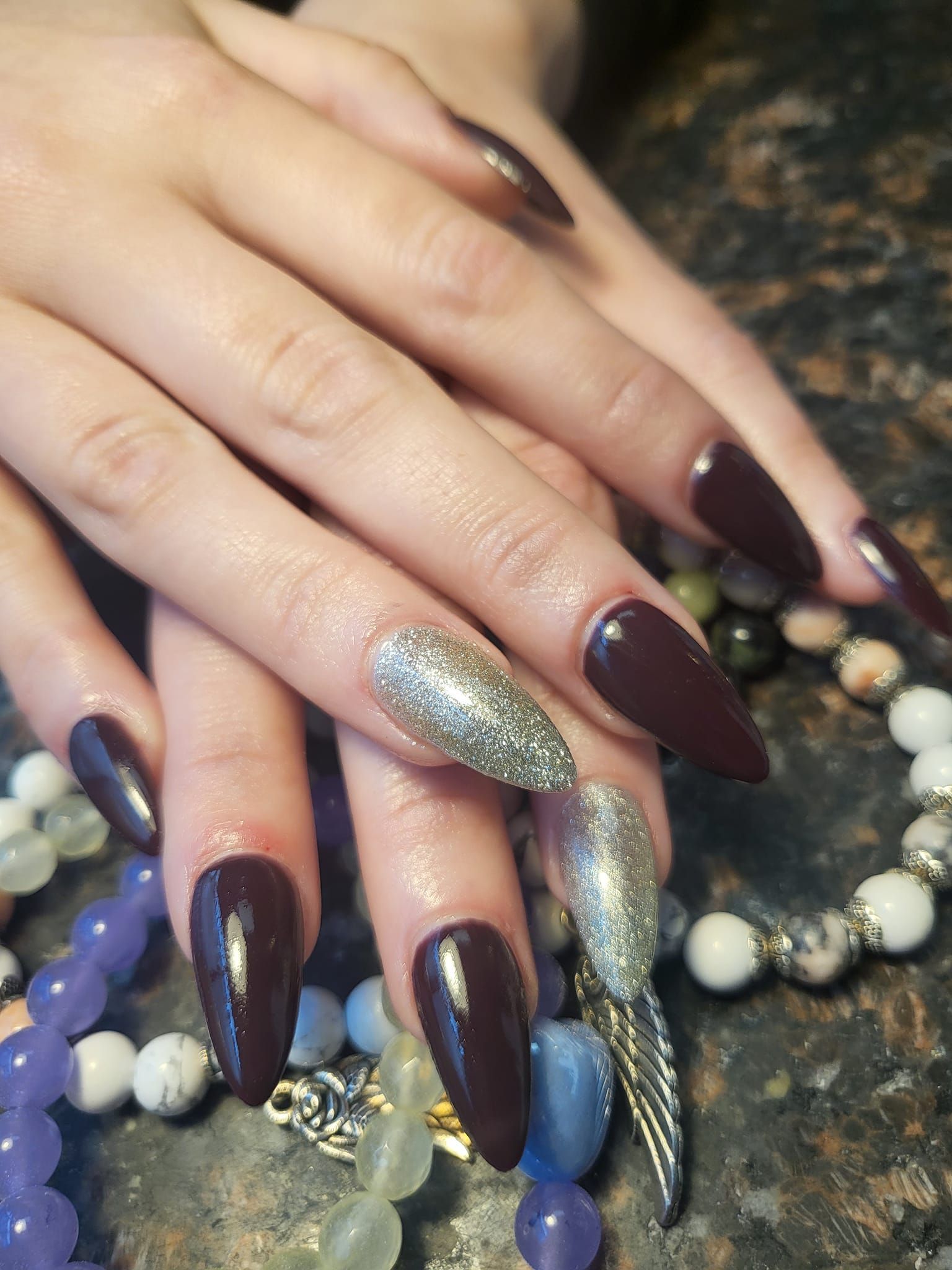 Hands with long, dark burgundy nails, one with silver glitter, resting on beaded jewelry.