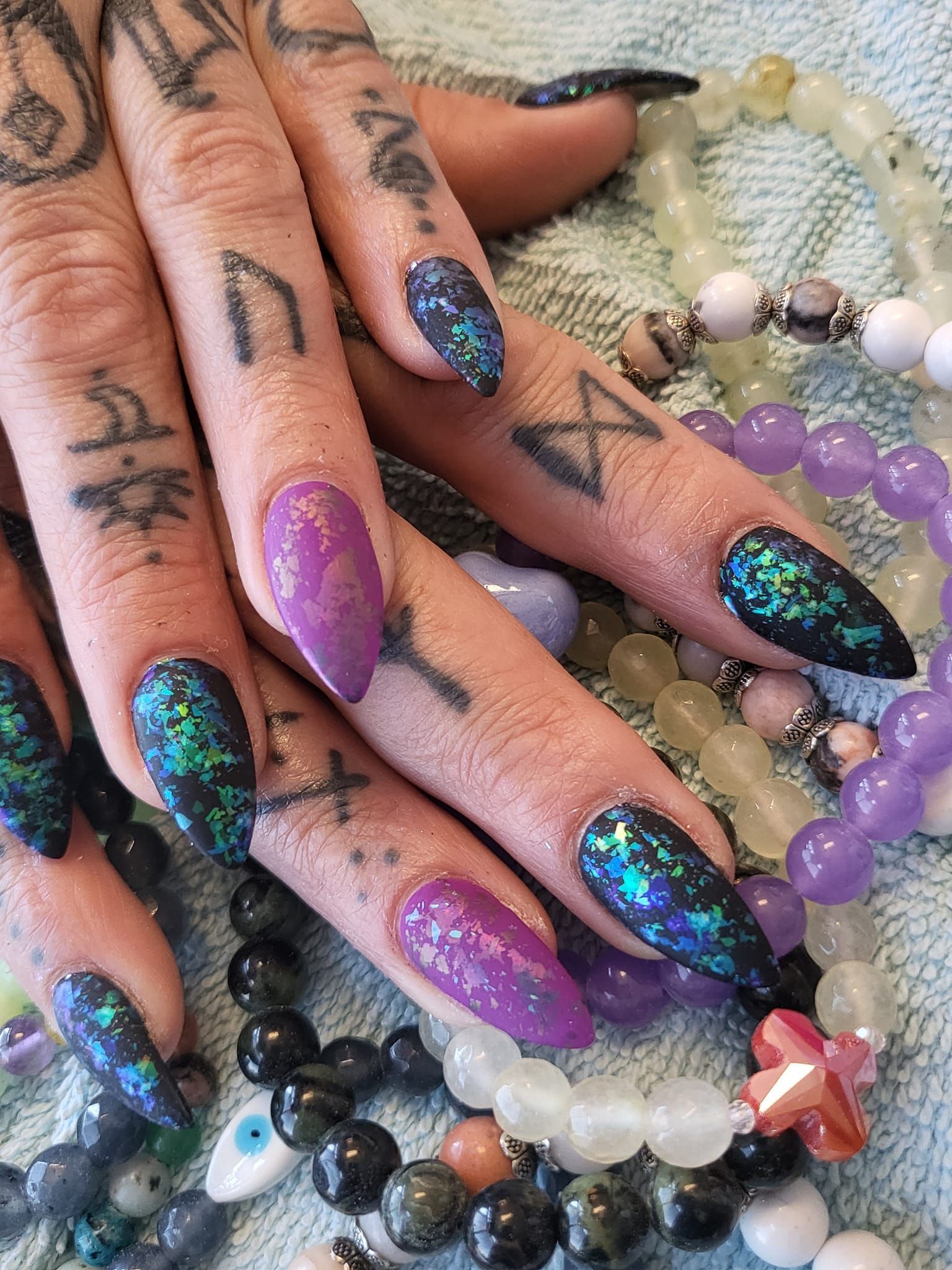 Hands with purple and teal glitter stiletto nails, surrounded by beaded bracelets.